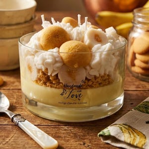 May include: A clear glass candle with a layered design resembling a banana pudding dessert. The candle features a yellow base, a crumbly layer, white frosting, and topped with cookies and banana slices. The candle has the text "Handmade with Love by Holligs Candle-Licious".