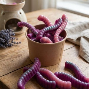 May include: A close-up of a brown paper cup filled with purple and pink worm-shaped wax melts. Several melts are scattered around the cup on a wooden surface. A ceramic oil warmer and lavender sprigs are in the background.