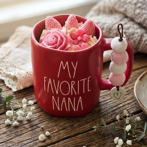 May include: A red ceramic mug with the text "MY FAVORITE NANA" in white. The mug is filled with pink floral decorations and topped with strawberry-shaped accents. A charm with pink and white pom-poms and a heart hangs from the handle.
