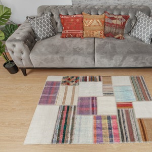 Puede incluir: Una alfombra blanca de patchwork con rayas y patrones de varios colores. La alfombra está colocada sobre un suelo de madera delante de un sofá gris con cojines.