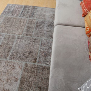 Może przedstawiać: Szary dywan patchworkowy w jasnoszarym, brązowym i białym kolorze. Dywan znajduje się przed szarym sofą z kolorowymi, wzorzystymi poduszkami.