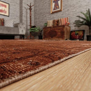 May include: A large rectangular area rug featuring a deep rust-brown color palette with intricate, darker brown geometric and floral patterns woven throughout the surface. The rug has a finished edge and is laid flat on a light-colored wooden floor.