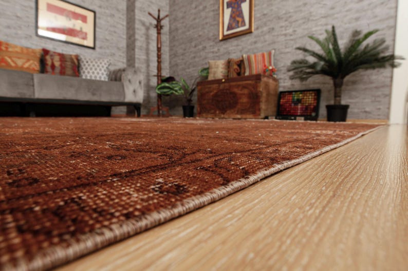 May include: A large rectangular area rug featuring a deep rust-brown color palette with intricate, darker brown geometric and floral patterns woven throughout the surface. The rug has a finished edge and is laid flat on a light-colored wooden floor.
