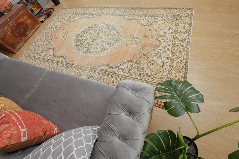 May include: An overhead shot of a large, rectangular, peach-colored rug with an intricate, traditional design. The rug is placed on a light wood floor, partially covered by a gray sofa and a potted plant with large green leaves.