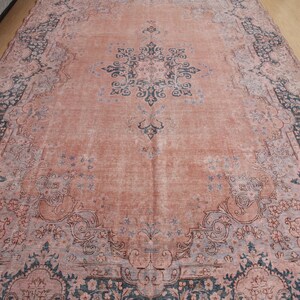 May include: A large rectangular area rug featuring a faded salmon pink field with a central dark blue floral medallion. The border displays intricate dark blue and light blue botanical patterns, creating a symmetrical design across the surface.