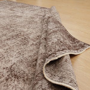 May include: A rolled-up, distressed-look rug with a faded brown and beige color palette. The rug's texture is visible, with a pattern of lines and subtle variations in tone. The edges of the rug are frayed, adding to its vintage aesthetic.