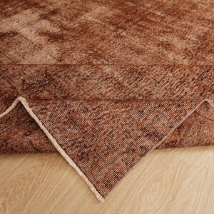 May include: A rectangular area rug featuring a distressed, weathered texture in shades of deep rust, copper, and brown. The rug is folded at one corner to reveal the woven underside, showcasing a consistent grid-like pattern and light-colored fringe.