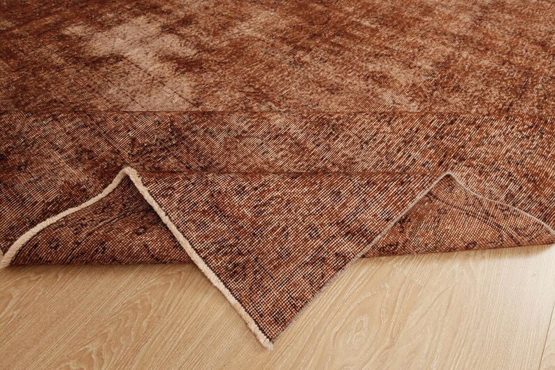 May include: A rectangular area rug featuring a distressed, weathered texture in shades of deep rust, copper, and brown. The rug is folded at one corner to reveal the woven underside, showcasing a consistent grid-like pattern and light-colored fringe.