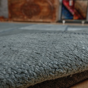 Może przedstawiać: Zbliżenie na szary, teksturowany dywan o miękkiej, puszystej powierzchni. Dywan ma subtelny wzór w jasnych i ciemnych odcieniach szarości.