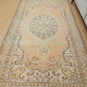 May include: A rectangular, peach-colored rug with an intricate, floral-inspired design. The rug features a central medallion and ornate borders in shades of cream, brown, and yellow. The rug is placed on a light-colored wooden floor.