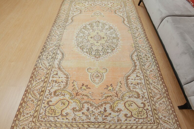 May include: A rectangular, peach-colored rug with an intricate, floral-inspired design. The rug features a central medallion and ornate borders in shades of cream, brown, and yellow. The rug is placed on a light-colored wooden floor.