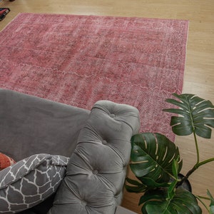 May include: A large, rectangular, faded rose-coloured rug with a detailed pattern. The rug is placed on a light-coloured wooden floor, partially covered by a grey sofa with decorative cushions and a large green plant.