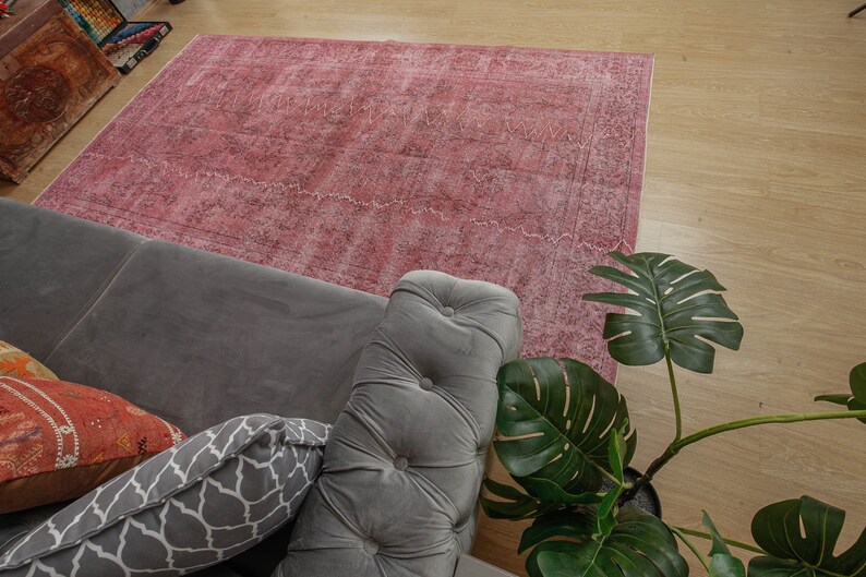 May include: A large, rectangular, faded rose-coloured rug with a detailed pattern. The rug is placed on a light-coloured wooden floor, partially covered by a grey sofa with decorative cushions and a large green plant.