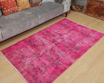 May include: A rectangular area rug featuring a vibrant hot pink base colour with a distressed, faded pattern in darker shades of purple and black. The rug has a low-pile texture and short white fringe detailing along the two shorter edges.