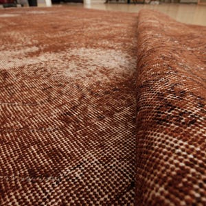 May include: A woven area rug featuring a deep rust-brown and cream color palette with a distressed, faded pattern. The rug is folded over to reveal the textured, knotted construction of the fabric, showing a dense pile and intricate weave details.