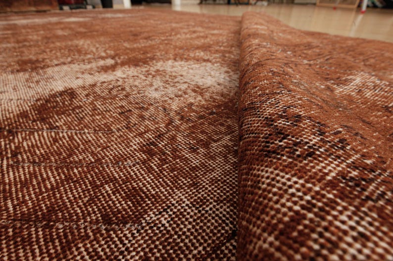 May include: A woven area rug featuring a deep rust-brown and cream color palette with a distressed, faded pattern. The rug is folded over to reveal the textured, knotted construction of the fabric, showing a dense pile and intricate weave details.