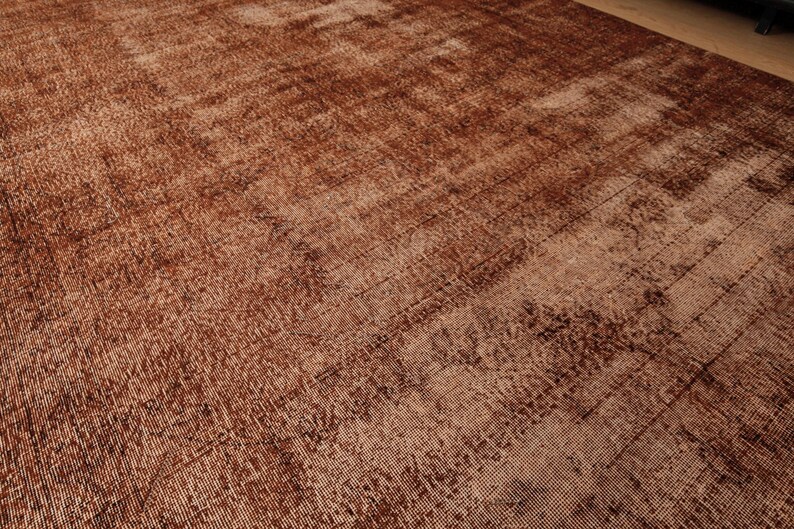 May include: A large rectangular area rug featuring a deep, monochromatic rust-brown color palette. The surface displays a distressed, weathered texture with visible woven fibers and subtle variations in tone across the entire flat textile.