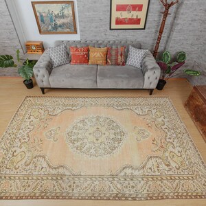 May include: A large, rectangular area rug with an intricate, traditional design. The rug features a central medallion in cream and peach tones, surrounded by a border with floral and geometric patterns in shades of brown, beige, and yellow. The rug is placed on a wooden floor.