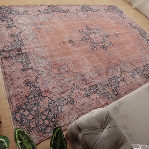 May include: A large square area rug featuring a faded terracotta orange center with a dark navy blue floral medallion. The border displays intricate dark navy blue and muted pink botanical patterns with a distressed, weathered appearance.
