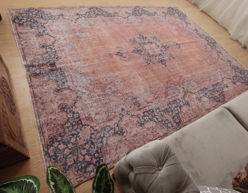 May include: A large square area rug featuring a faded terracotta orange center with a dark navy blue floral medallion. The border displays intricate dark navy blue and muted pink botanical patterns with a distressed, weathered appearance.