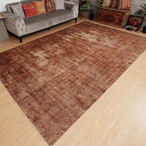 May include: A large rectangular area rug featuring a distressed, weathered texture in various shades of deep brown, copper, and muted tan. The rug displays a faded, mottled pattern across its surface, with lighter beige highlights creating a worn appearance.