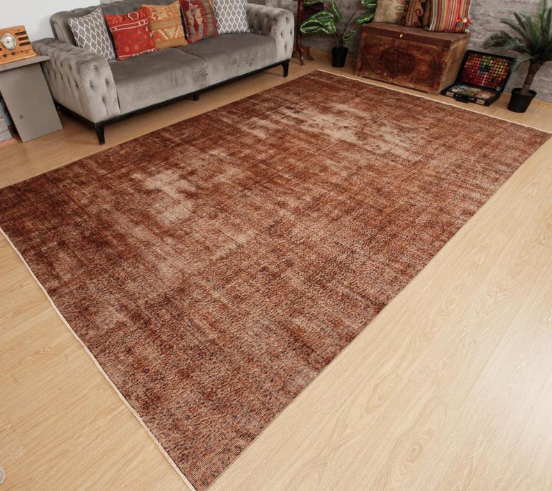 May include: A large rectangular area rug featuring a distressed, weathered texture in various shades of deep brown, copper, and muted tan. The rug displays a faded, mottled pattern across its surface, with lighter beige highlights creating a worn appearance.
