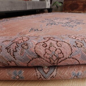 May include: A large area rug featuring a faded salmon pink base color with intricate floral and geometric patterns in muted blue and grey tones. The rug is partially folded over, revealing the woven texture and the detailed design on the surface.