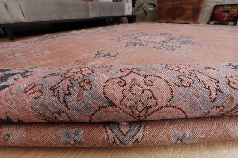 May include: A large area rug featuring a faded salmon pink base color with intricate floral and geometric patterns in muted blue and grey tones. The rug is partially folded over, revealing the woven texture and the detailed design on the surface.