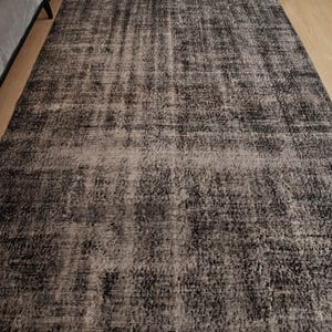 May include: A rectangular area rug featuring a distressed, mottled pattern in shades of charcoal grey, black, and muted beige. The surface displays a textured, weathered appearance with varying colour density across the woven fabric construction.