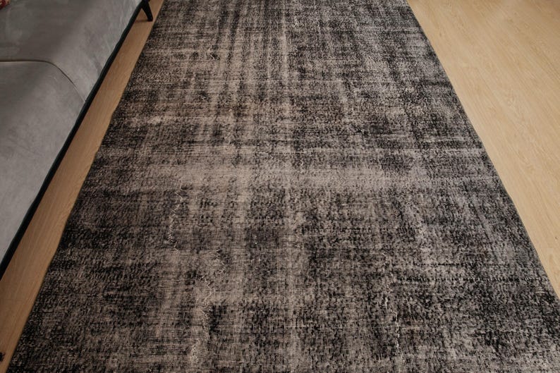 May include: A rectangular area rug featuring a distressed, mottled pattern in shades of charcoal grey, black, and muted beige. The surface displays a textured, weathered appearance with varying colour density across the woven fabric construction.