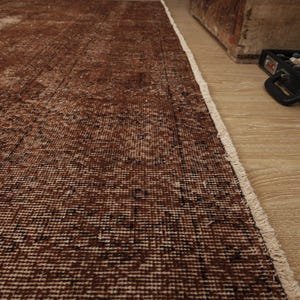 May include: A rectangular area rug featuring a distressed, deep brown and black woven pattern with a visible white fringe along the edge. The rug displays a textured, weathered appearance with variations in color density across the surface.