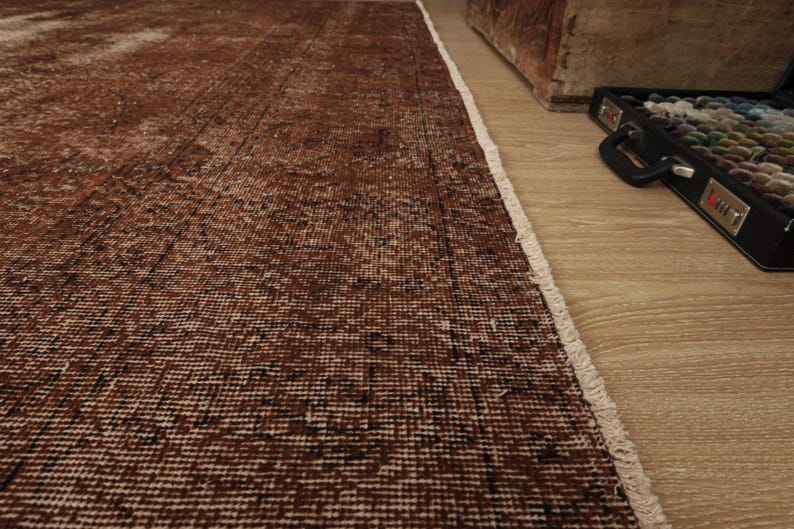May include: A rectangular area rug featuring a distressed, deep brown and black woven pattern with a visible white fringe along the edge. The rug displays a textured, weathered appearance with variations in color density across the surface.