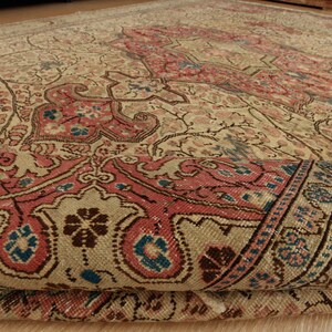 Pu&ograve; includere: Un tappeto antico con un motivo floreale dettagliato nei toni del beige, rosa e blu. Il design del tappeto presenta un medaglione centrale e un bordo intricato, con un bordo arrotolato che rivela lo spessore e la trama.