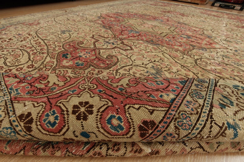 Pu&ograve; includere: Un tappeto antico con un motivo floreale dettagliato nei toni del beige, rosa e blu. Il design del tappeto presenta un medaglione centrale e un bordo intricato, con un bordo arrotolato che rivela lo spessore e la trama.