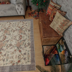 May include: A beige rug with a floral pattern and a gray border. The rug is in a living room with a gray sofa, a wooden chest, and a variety of pillows.