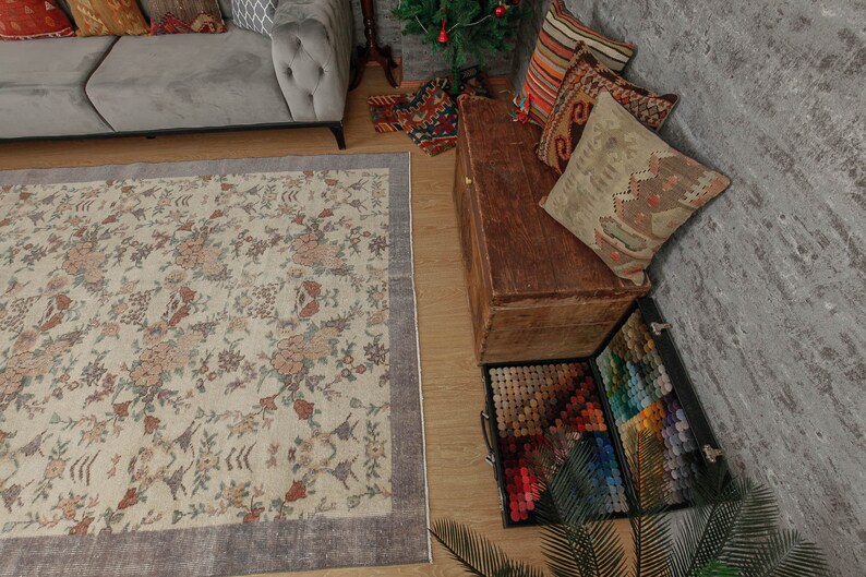 May include: A beige rug with a floral pattern and a gray border. The rug is in a living room with a gray sofa, a wooden chest, and a variety of pillows.