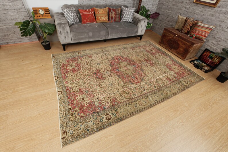 Pu&ograve; includere: Un tappeto beige antico con un motivo a medaglione centrale in rosso e verde. Il tappeto presenta intricati motivi floreali e un bordo con finiture verde scuro e blu. Il tappeto &egrave; posato su un pavimento in legno chiaro.