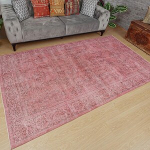 May include: A large, rectangular, distressed pink rug with a detailed border and faded floral patterns. The rug is placed on a light wood floor. The rug's dimensions are visible, with a gray sofa in the background.
