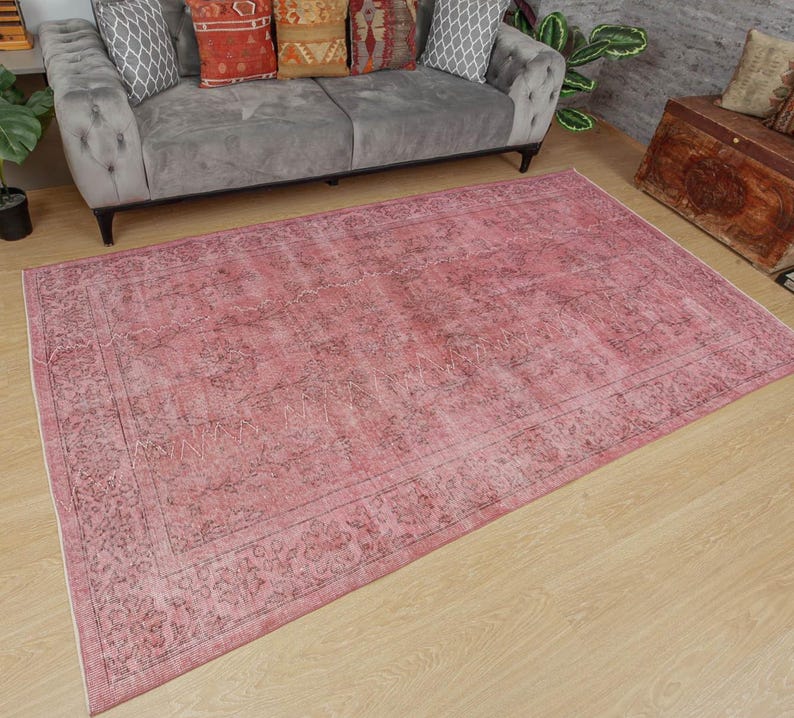 May include: A large, rectangular, distressed pink rug with a detailed border and faded floral patterns. The rug is placed on a light wood floor. The rug's dimensions are visible, with a gray sofa in the background.
