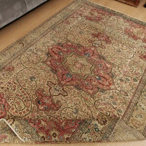 Pu&ograve; includere: Un tappeto beige antico con un medaglione centrale rosso, blu e oro. Il tappeto presenta un intricato motivo floreale e un bordo dettagliato. Il tappeto &egrave; parzialmente piegato, rivelando il suo rovescio.