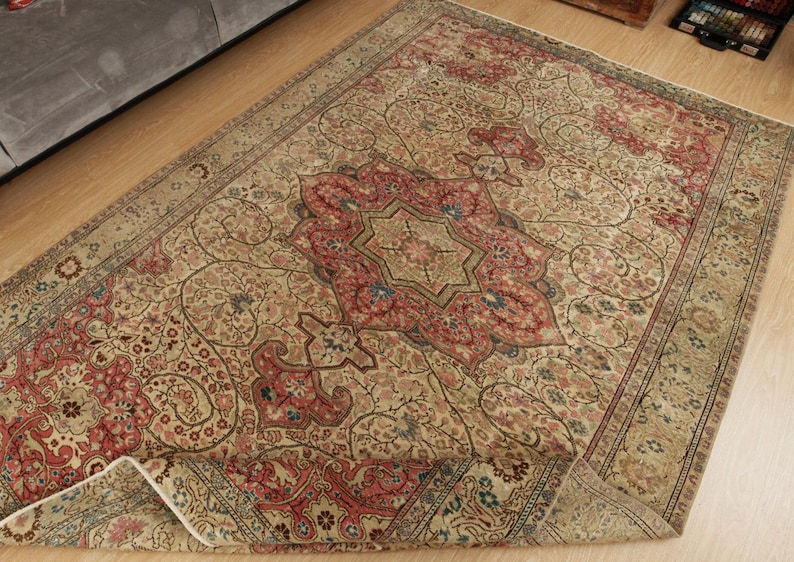 Pu&ograve; includere: Un tappeto beige antico con un medaglione centrale rosso, blu e oro. Il tappeto presenta un intricato motivo floreale e un bordo dettagliato. Il tappeto &egrave; parzialmente piegato, rivelando il suo rovescio.