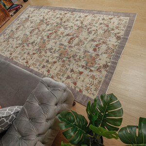 May include: A cream-colored rug with a floral pattern in shades of brown, pink, and purple. The rug is laid out on a wooden floor and a gray sofa with pillows is in the foreground.