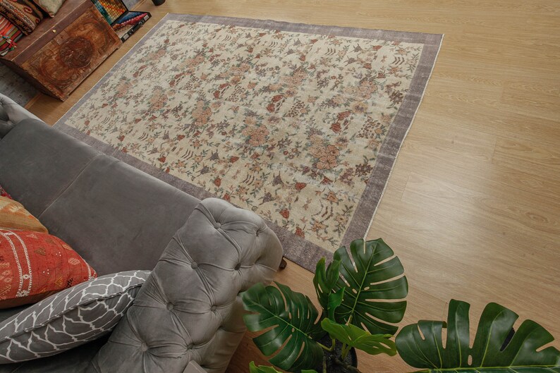 May include: A cream-colored rug with a floral pattern in shades of brown, pink, and purple. The rug is laid out on a wooden floor and a gray sofa with pillows is in the foreground.