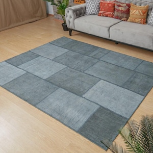 May include: A rectangular, patchwork-style rug in shades of gray and blue. The rug is composed of various rectangular pieces sewn together, creating a geometric pattern. The rug is placed on a light-colored wooden floor.