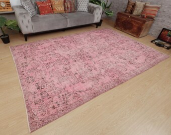 May include: A rectangular area rug featuring a faded pink base color with intricate, dark-toned floral and vine patterns throughout. The rug has a distinct border design and a low-pile texture, covering a large portion of the light-colored wooden floor.