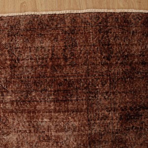 May include: A rectangular area rug featuring a deep rust-brown color palette with intricate, faded dark brown patterns woven throughout the fabric. The rug has a visible, light-colored fringe along the top edge, showcasing a dense, textured weave.