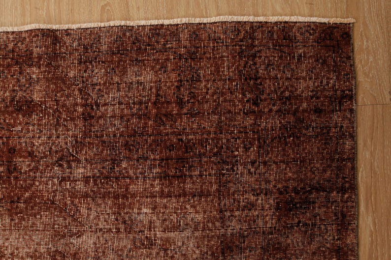 May include: A rectangular area rug featuring a deep rust-brown color palette with intricate, faded dark brown patterns woven throughout the fabric. The rug has a visible, light-colored fringe along the top edge, showcasing a dense, textured weave.