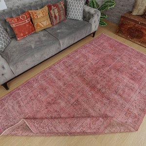 May include: A large, rectangular rug in shades of pink and faded black, with a distressed appearance. The rug has an intricate, floral-inspired pattern and is partially rolled up. It is placed on a light-coloured wooden floor.