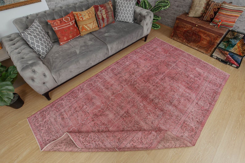 May include: A large, rectangular rug in shades of pink and faded black, with a distressed appearance. The rug has an intricate, floral-inspired pattern and is partially rolled up. It is placed on a light-coloured wooden floor.