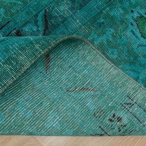 May include: Close-up of the back side of a teal blue rug with a woven pattern. The rug is folded in half, showing the texture of the fabric.